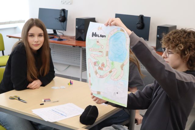 Na zdjęciu znajdują się dwie dziewczyny przy stoliku, dziewczyna po prawej podnosi i pokazuje wykonaną przez ilustrację przedstawiając planszę do gry z jeziorem, pałacem i napisem Malbork. Na stoliku leżą kartki, kredki, nożyczki i piórnik. Warsztaty odbywają się w sali komputerowej.