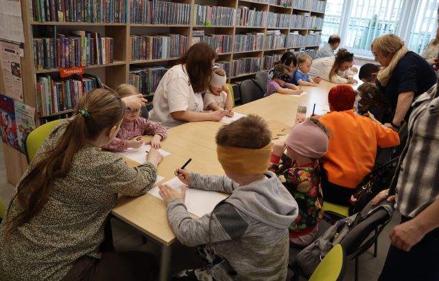 Na zdjęciu znajdują się dzieci i trójka dorosłych przy złączonych stołach. Część dzieci ma zasłoniete oczy i rysuje na kartkach. W tle znajdują się półki z książkami.