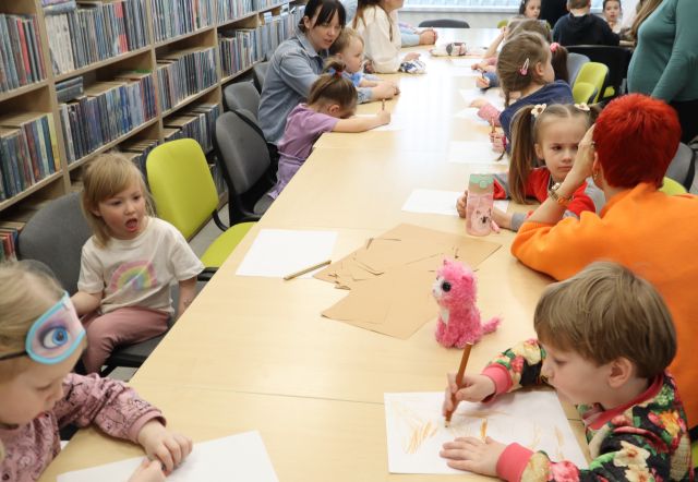 Na zdjęciu znajdują się dzieci siedzące przy złączonych stołach i rysują na kartkach z zasłonietymi oczami. Na stole leża jasnobrązowe kartki. W tle znajdują się półki z książkami.