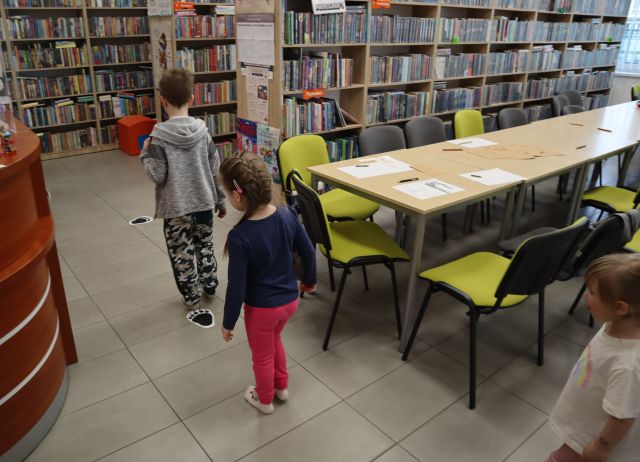 Na zdjęciu znajdują się dzieci, które chodzą po śladach niedźwiedzia przy stołach i półkach z książkami.