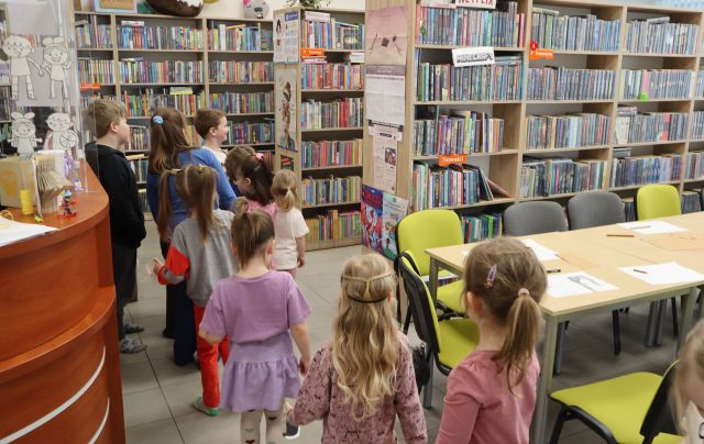 Na zdjęciu znajdują się dzieci jeden za drugim między stolikami, półkami z książkami a biurkiem.