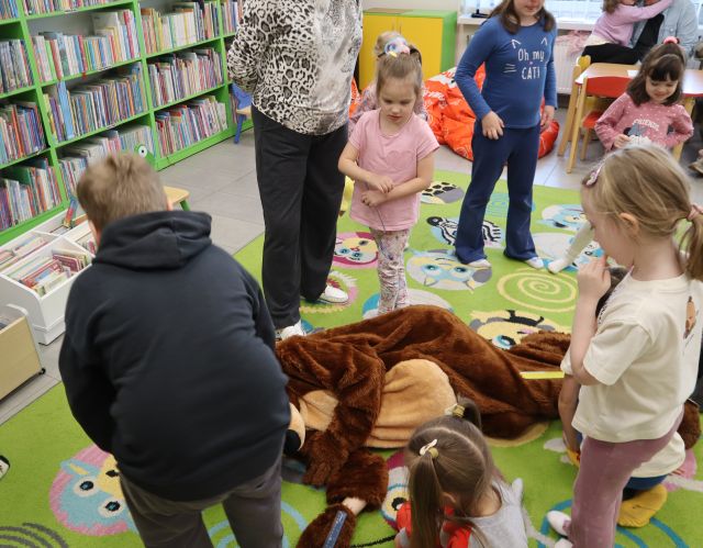 Na zdjęciu znajduje się leżący na dywanie człowiek w przebraniu niedźwiedzia, a dookoła niego stoją dzieci, jeden chłopiec się nachyla do niedźwiedzia. W tle znajdują się półki z książkami.
