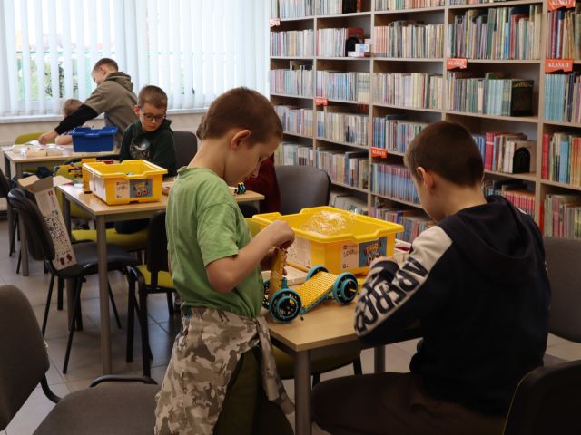 Na zdjęciu znajduje się szóstka chłopców przy trzech stolikach, przy których w parach układają klocki LEGO. W tle znajdują się półki z książkami.