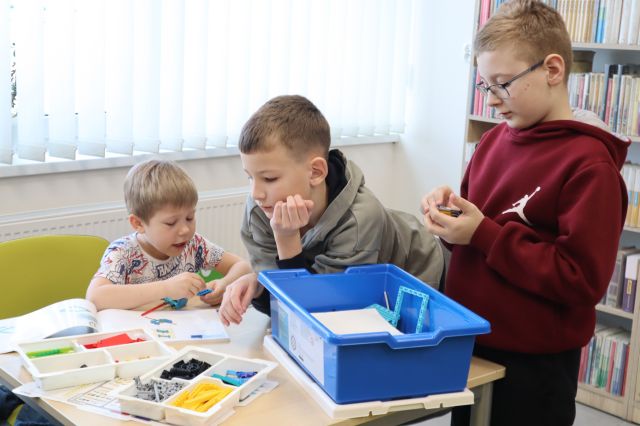 Na zdjęciu znajduje się trójka chłopców przy stoliku, jeden stoi, a dwójka siedzi i układają klocki LEGO, które leżą na stoliku i w niebieskim pudełku. W tle znajdują się okna i fragment półek z książkami po prawej stronie.