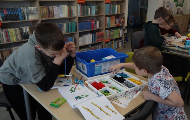 Na zdjęciu znajduje się dwójka chłopców składająca wspólnie klocki LEGO, które leżą na stole przed nimi wraz z instrukcją i niebieskim pudełkiem. W tle znajdują się półki z książkami oraz dwóch chłopców przy stoliku obok.