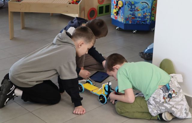 Na zdjęciu znajduje się trójka chłopców na podłodze i nachylają się nad zbudowanym z klocków LEGO samochodem, jeden z nich ma przed sobą tablet. W tle znajdują się meble.