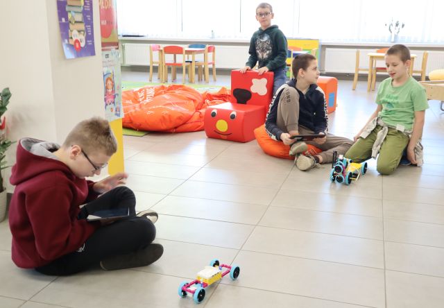 Na zdjęciu znajduje się czwórka chłopców, jeden siedzący na podłodze po lewej stronie zdjęcia i trzyma przed sobą tablet, na przeciwko niego siedzi dwóch chłopców, jeden z nich również trzyma tablet, a obok stoi chłopiec przy pociągu z puf. Między chłopcami na podłodze znajdują się dw robotyczne samochody z klocków LEGO. W tle znajdują się czerwone pufy oraz stoliki z krzesełkami.