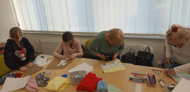Na zdjęciu znajdują się dwie kobiety oraz dwie dziewczyny przy załączonych stolikach i odrysowują na materiale szablon. Przed nimi na stoliku znajdują się kolorów materiały, a za nimi okna.