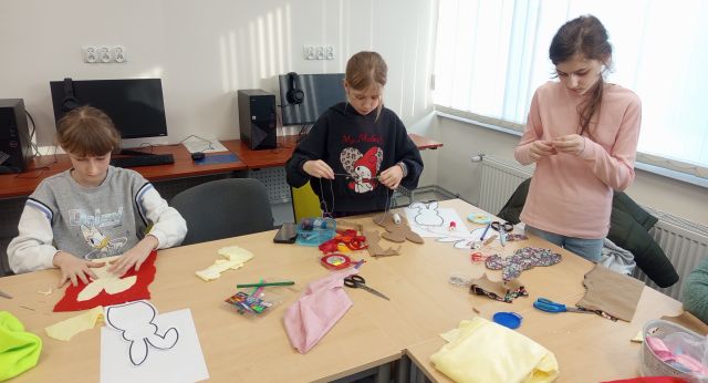 Na zdjęciu znajdują się trzy dziewczyny szyjce maskotkę królika że znajdujących się przed nimi na załączonych stolikach materiałów. Warsztaty odbywają się w sali komputerowej.