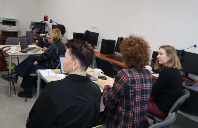 Na zdjęciu znajdują się trzy kobiety przy stoliku, na którym znajduje się laptop, przed nimi pry stoliku siedzą dwie kobiety. Warsztaty odbywają się w sali komputerowej.