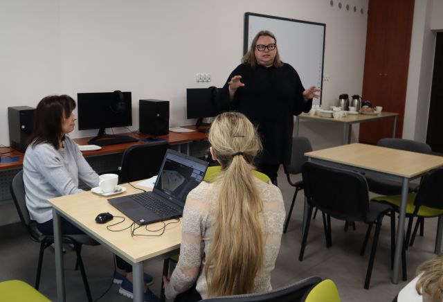 Na zdjęciu znajduje się stojąca i gestykulująca kobieta pomiędzy dwoma stolikami, przy stoliku po lewej siedzą dwie kobiety, a na stoliku leży laptop, drugi stolik jest wolny. Warsztaty odbywają się w sali komputerowej.