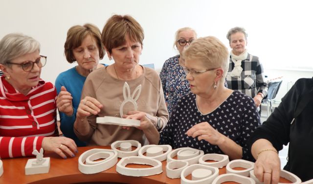Na zdjęciu znajdują się kobiety, które wybierają gipsowe ozdoby takie jak króliczki i jajka z drewnianego blatu. Jedna z kobiet trzyma w ręce wzór królika. W tle znajdują się zasłonięte okna.