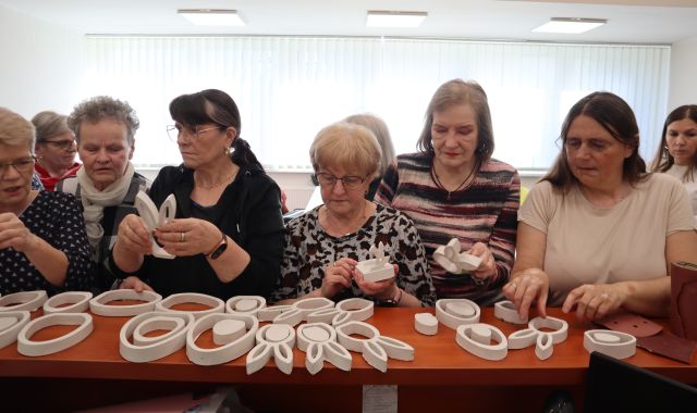 Na zdjęciu znajdują się kobiety wybierające z blatu wielkanocne ozdoby z gipsu, takie jak jajka i króliki. W tle znajdują się zasłonięte okna.