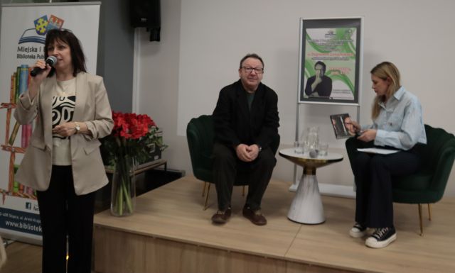 Na zdjęciu znajduje się mężczyzna oraz kobieta na fotelach na scenie, między nimi stoi stolik oraz plakat promujący spotkanie. Koło sceny stoi kobieta z mikrofonem przy ustach, a za nią w tle znajduje się baner z logiem biblioteki i z książkami.