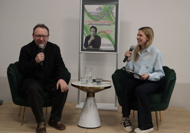 Na zdjęciu znajduje się mężczyzna oraz kobieta, którzy siedzą na scenie, w ręce trzymają mikrofon. Pomiędzy nimi stoi stolik z wodą, a za nim plakat promujący spotkanie.