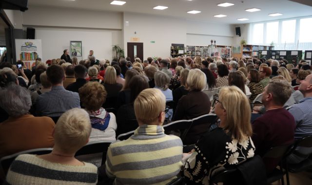 Na zdjęciu znajduje się około dwustu ludzi, którzy siedzą na holu biblioteki i skierowani są w stronę sceny, na której siedzi mężczyzna oraz kobieta.