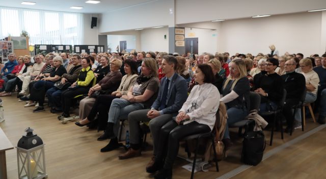 Na zdjęciu znajduje się około dwustu osób siedzących na holu biblioteki. W tle znajdują się okna.