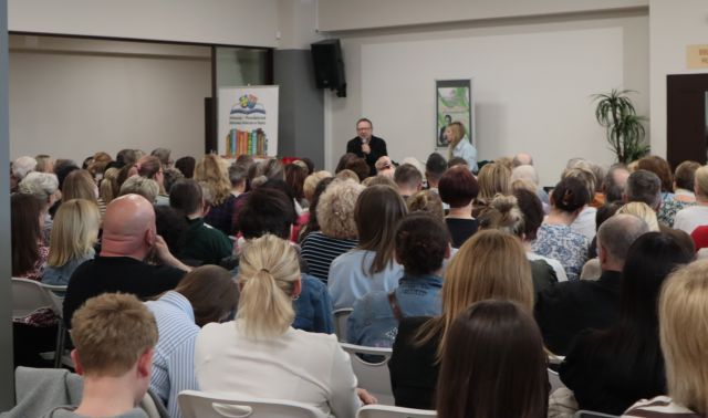 Na zdjęciu znajdują się ludzie, którzy siedzą na holu biblioteki i skierowani są w stronę mężczyzny oraz kobiety, którzy siedzą na scenie, obok nich stoi plakat promujący spotkanie, a przy scenie baner z logiem biblioteki.