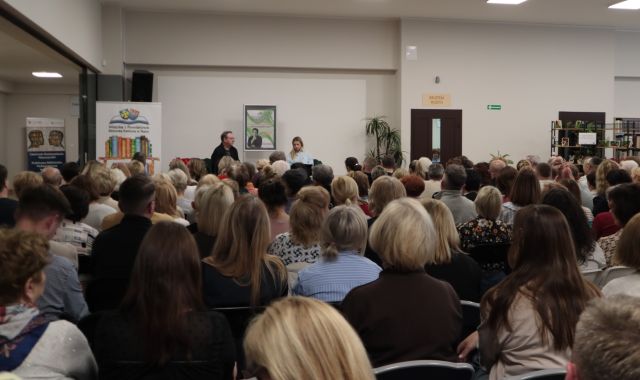 Na zdjęciu znajdują się ludzie siedzący na holu biblioteki, skierowani w stronę sceny, na której znajduje się mężczyzna oraz kobieta, za nimi stoi plakat promujący spotkanie, a po lewej stronie przy scenie baner z książkami i logiem biblioteki.