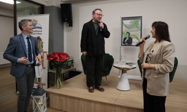 Na zdjęciu znajduje się mężczyzna stojący na scenie, obok niego stoi stolik oraz plakat promujący spotkanie. Po prawej stronie pod sceną stoi kobieta z mikrofonem, a po lewej stronie mężczyzna trzymający w ręce mała torebkę prezentową, za mężczyzną stoi baner z książkami i logiem biblioteki oraz czerwone kwiaty.