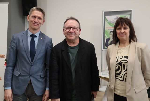 Na zdjęciu znajduje się dwoje mężczyzn oraz kobieta, którzy stoją wspólnie do zdjęcia. Za kobietą znajduje się plakat promujący spotkanie.