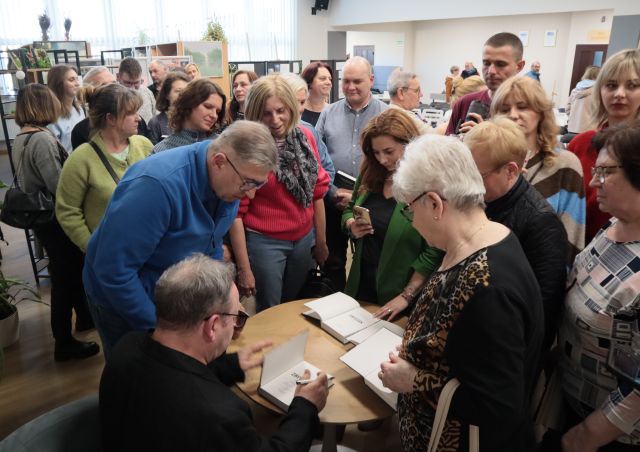 Na zdjęciu znajduje się mężczyzna podpisujący przy stoliku książki, a wokół niego znajduje siębardzo dużo ludzi.