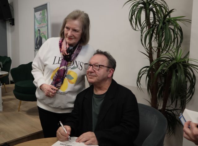 Na zdjęciu znajduje się mężczyzna siedzący przy stoliku, a obok niego stoi kobieta i uśmiechają się do wspólnego zdjęcia. W tle znajduje się scena z fotelami i plakatem promującym spotkanie. A po prawej stronie zdjęcia stoi palma.