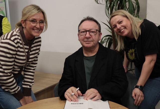 Na zdjęciu znajduje się mężczyzna siedzący przy stoliku, a po jego lewej i prawej stronie nachylają się kobiety do wspólnego zdjęcia. Po prawej stronie za kobietą znajduje się palma.