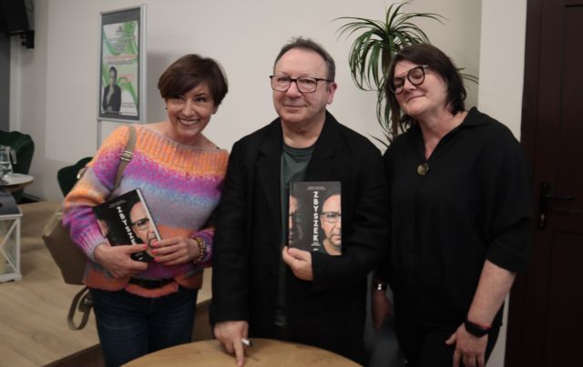 Na zdjęciu znajduje się mężczyzna oraz dwie kobiety, którzy stoją wspólnie do zdjęcia. Mężczyzna oraz kobieta stojąca po lewej stronie trzymają książki. W tle znajduje się scena oraz palma.