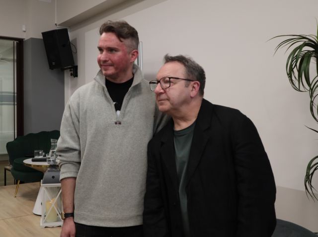 Na zdjęciu znajduje się dwóch mężczyzn stojących wspólnie do zdjęcia, za nimi znajduje się scena, a po prawej stronie zdjęcia znajduje się kawałek palmy.