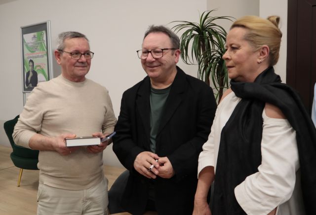 Na zdjęciu znajduje się dwóch mężczyzn, jeden z nich trzyma książkę oraz kobieta i stoją wspólnie do zdjęcia. W tle znajduje się scena z plakatem promującym spotkanie oraz palma po prawej stronie.