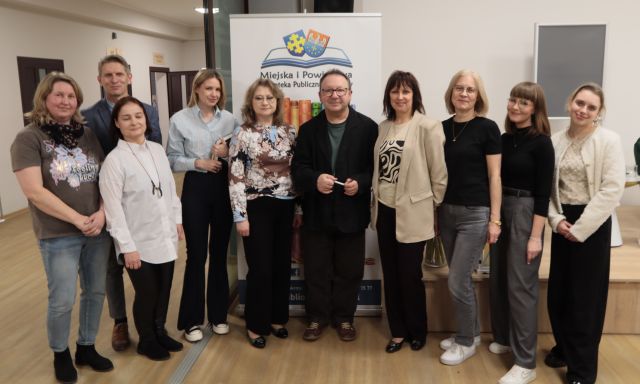Na zdjęciu się dwóch mężczyzn oraz osiem kobiet stojących wspólnie do zdjęcia na holu biblioteki, za nimi stoi baner z logiem biblioteki.