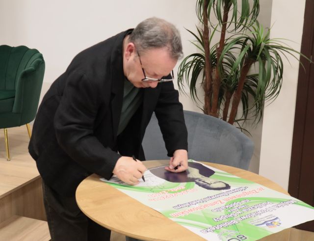 Na zdjęciu znajduje się mężczyzna nachylający się nad stolikiem i podpisuje plakat promujący spotkanie. Za mężczyzną znajduje się palma oraz scena z zielonym fotelem.