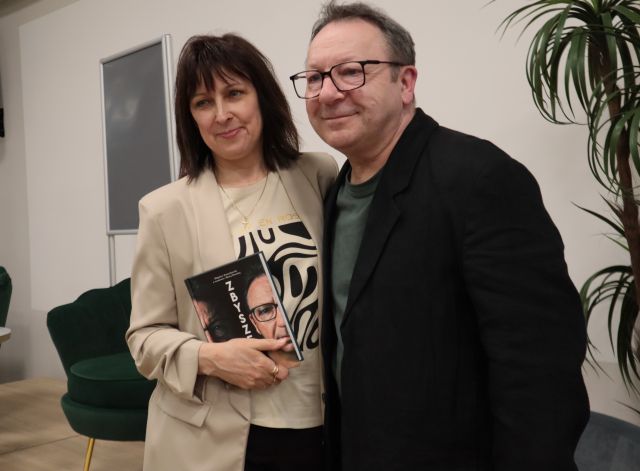 Na zdjęciu znajduje się kobieta, trzymająca w ręce książkę i stoi do zdjęcia z mężczyzną. W tle znajduje się scena z fotelami i plakatem, oraz palma po prawej stronie.