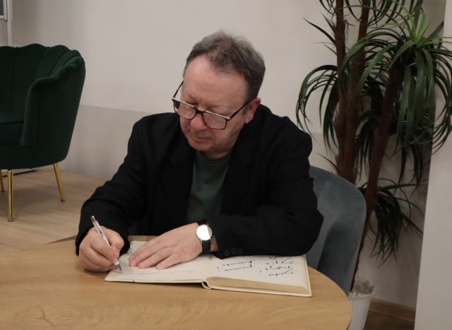 Na zdjęciu znajduje się mężczyzna siedzący przy stoliku i wpisuje się do księgi, z nim po prawej stronie stoi palma, a po lewej stronie fotel na scenie.