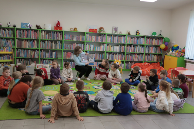Na zdjęciu znajduje się grupa przedszkolaków siedząca w kółku na dywanie, pomiędzy nimi siedzi kobieta i pokazuje im ilustracje w książce. w tle znajdują się półki z książkami.