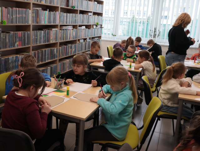 Na zdjęciu znajdują się przedszkolaki przy stolikach i odrysowują na kartkach słoiki. W tle znajdują się okna oraz półki z książkami.