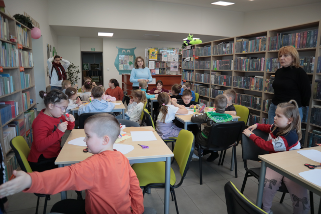 Na zdjęciu znajdują się przedszkolaki przy stolikach i rysują, przed nimi stoi kobieta. Po lewej i prawej stronie znajdują się półki z książkami.