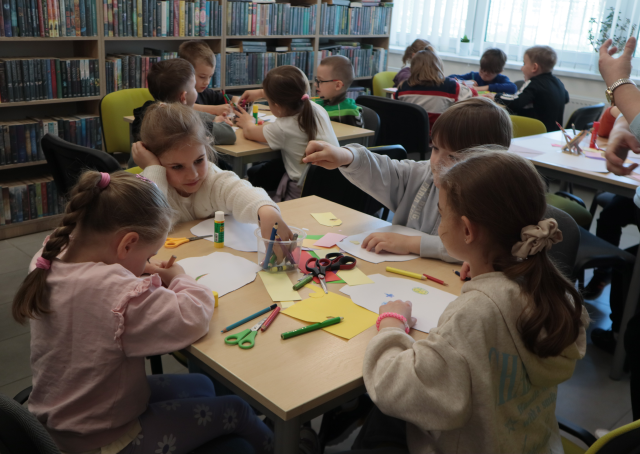 Na zdjęciu znajdują się przedszkolaki przy stoliku i kolorują papierowe słoiczki. W tle znajdują się półki z książkami.
