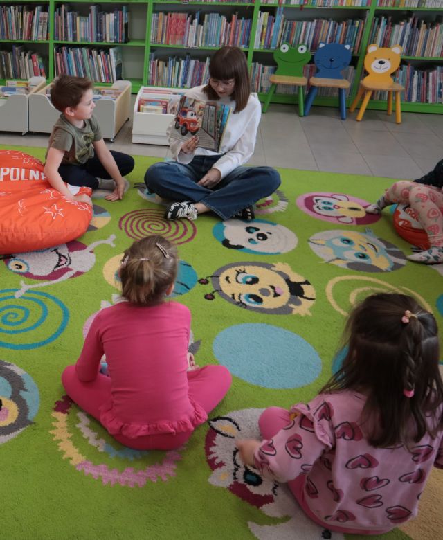 Na zdjęciu znajdują się dzieci wraz z kobietą na dywanie, która pokazuje im ilustracje książki. W tle znajdują się półki z książkami oraz trzy małe krzesełka.