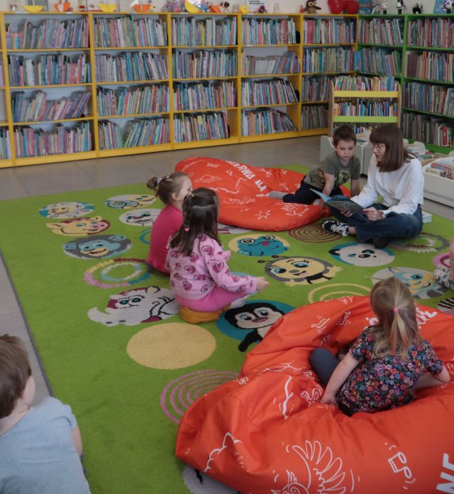 Na zdjęciu znajduje się dzieci na dywanie wraz z kobietą, która czyta im książkę. Na dywanie znajdują się również czerwone pufy, na których siedzą dwie osoby. W tle znajdują się półki z książkami.