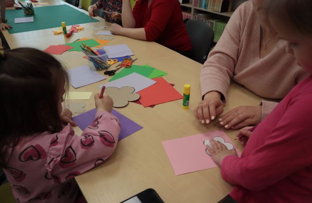 Na zdjęciu znajduje się stół wraz z kolorowymi kartkami przy którym siedzą dzieci wraz z rodzicami i odrysowują kwiatki.