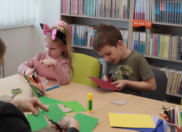 Na zdjęciu znajduje się dwójka dzieci przy złączonych stołach i wycinają z kolorowych kartek kwiatki. Za nimi w tle znajdują się półki z książkami.