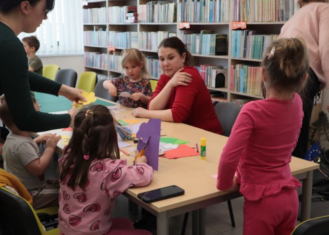 Na zdjęciu znajdują się dzieci wraz z rodzicami przy złączonych stołach przy których wspólnie wycinają kwiatki. W tle znajdują się półki z książkami