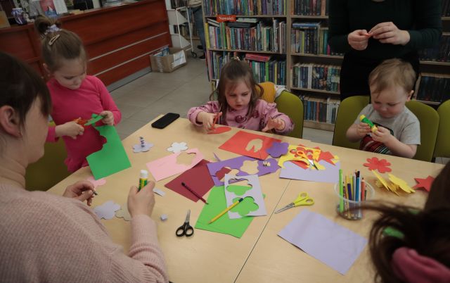 Na zdjęciu znajduje się trójka dzieci przy złączonych stołach wraz z rodzicami. Na stole znajdują się kolorowe kartki, z których dzieci wycinają kolorowe kwiatki. W tle znajdują się półki z książkami. 