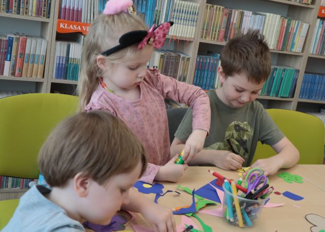 Na zdjęciu znajduje się trójka dzieci przy stole, które wycina i klei kolorowe kwiatki. W tle znajdują się półki z książkami.