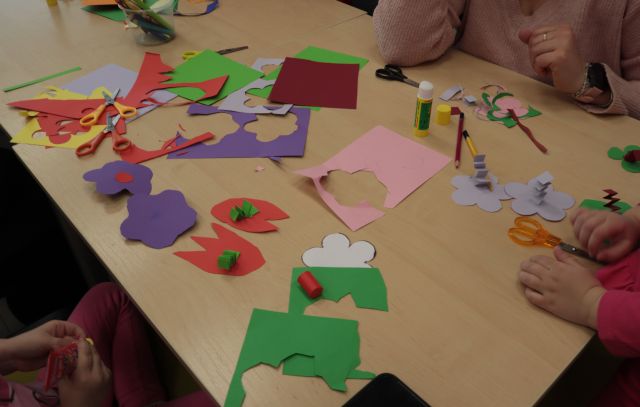 Na zdjęciu znajdują się złączone stoły z kolorowymi kartkami i wyciętymi kwiatkami, przy stoliku siedzą dwie osoby, które nie zostały uchwycone na zdjęciu całe.