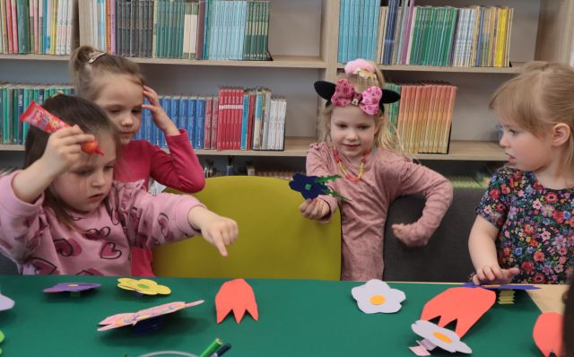 Na zdjęciu znajduje się czwórka dziewczynek przed złączonymi stołami na których leży zielony brystol i przyklejają do niego kolorowe kwiatki. Na środku między dziewczynkami stoi krzesło, a w tle półki z książkami.