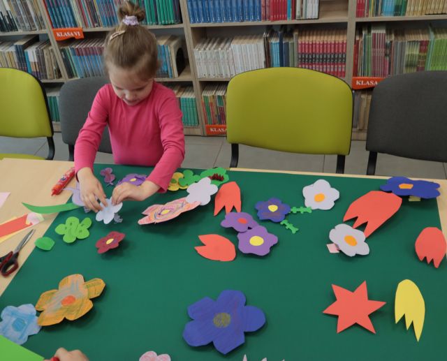 Na zdjęciu, po lewej znajduje się dziewczynka przyklejająca kolorowe kwiatki do brystolu lżącego na złączonych stołach. Przy dziewczynce znajdują się krzesła, a za nią półki z książkami.