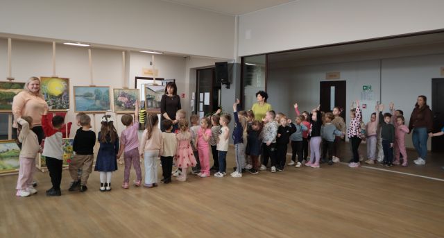 Na zdjęciu znajdują się przedszkolaki stojące w parach, część z nich podnosi rękę do góry, razem z nimi stoją cztery kobiety. Grupa stoi na holu biblioteki, za nimi stoją obrazy oraz są otwarte drzwi.